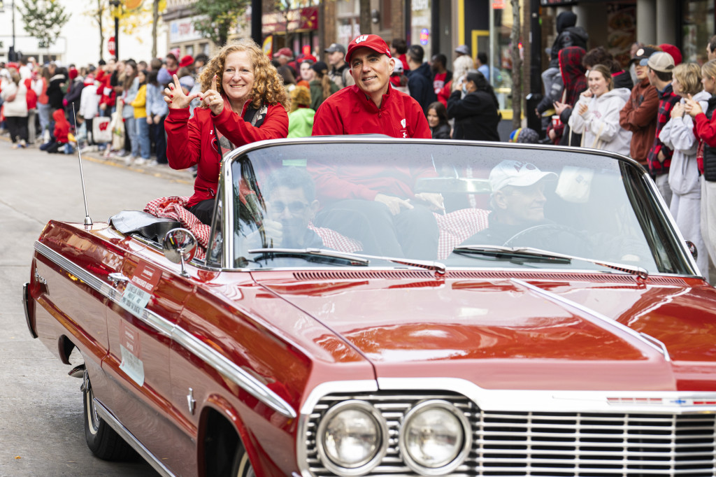 Seated on the back of a red, 1964 convertible, Jennifer Mnookin makes a W with her hands as she and Joshua Foa Dienstag smile at the crowd.