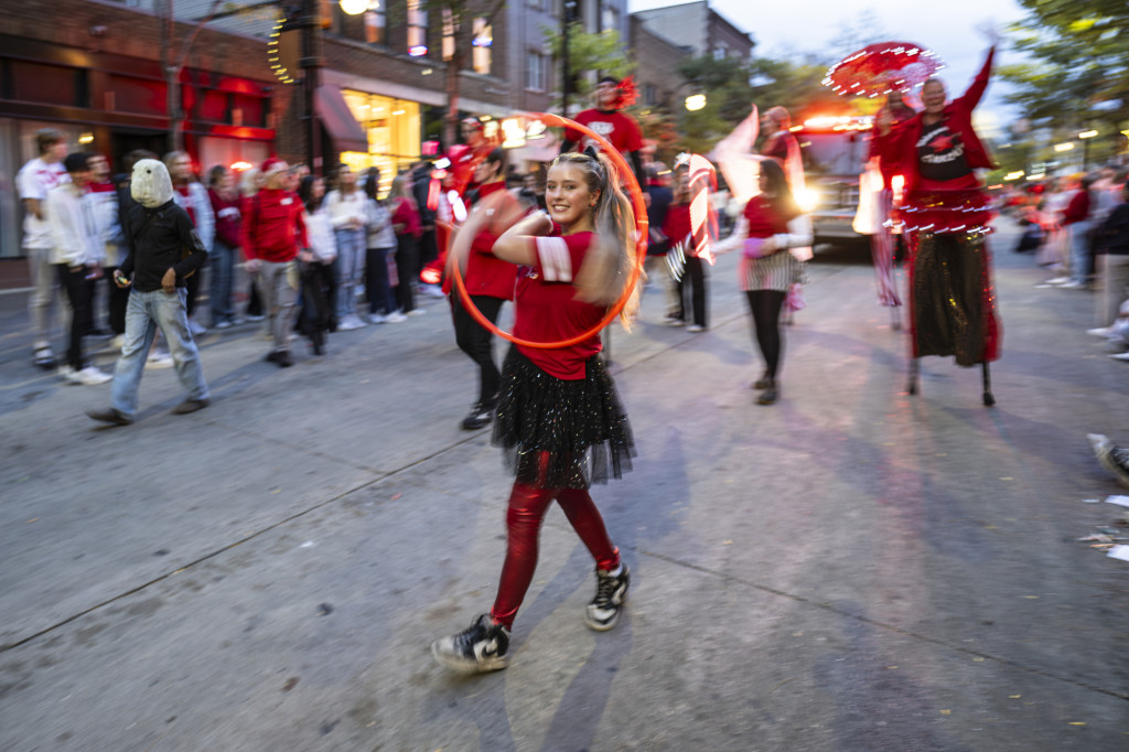 A woman wearing a red top and a black skirt waves a red, sparkling ribbon in a circle as she leads a circus troupe down the parade route.