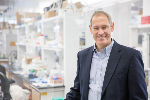 A man wearing a sports coat and an Oxford button-down shirt stands in a brightly lit chemistry lab.