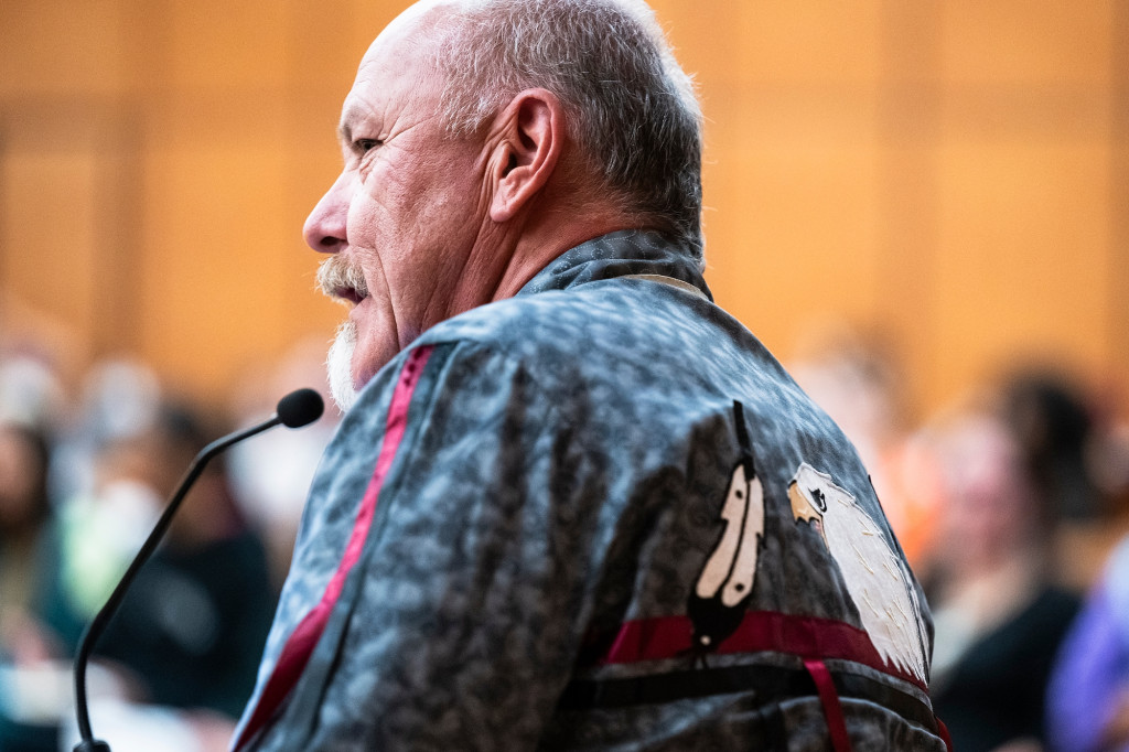 A profile view of a man wearing a blue shirt adorned with a bald eagle and eagle feathers speaks into a microphone.