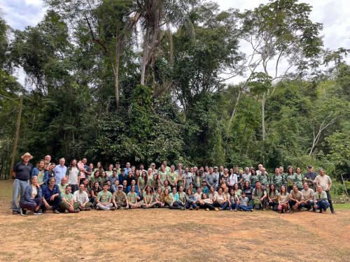 A group of nearly 100 people sit and stand in rows, smiling at the camera with a forest backdrop behind them.