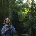 A woman holds binoculars away from her face as she looks up into a dense tree canopy.