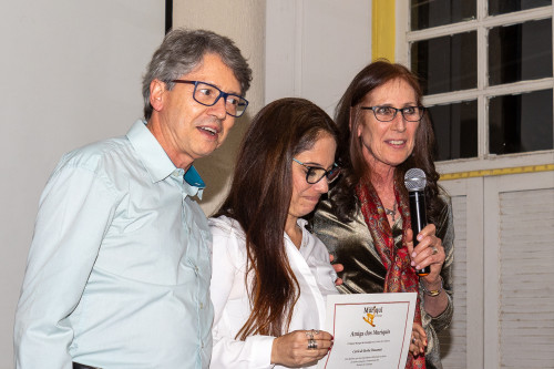 A man and a woman stand on either side of the woman between them. They're smiling and heaping praise on the woman in the center, who is looking down and smiling.