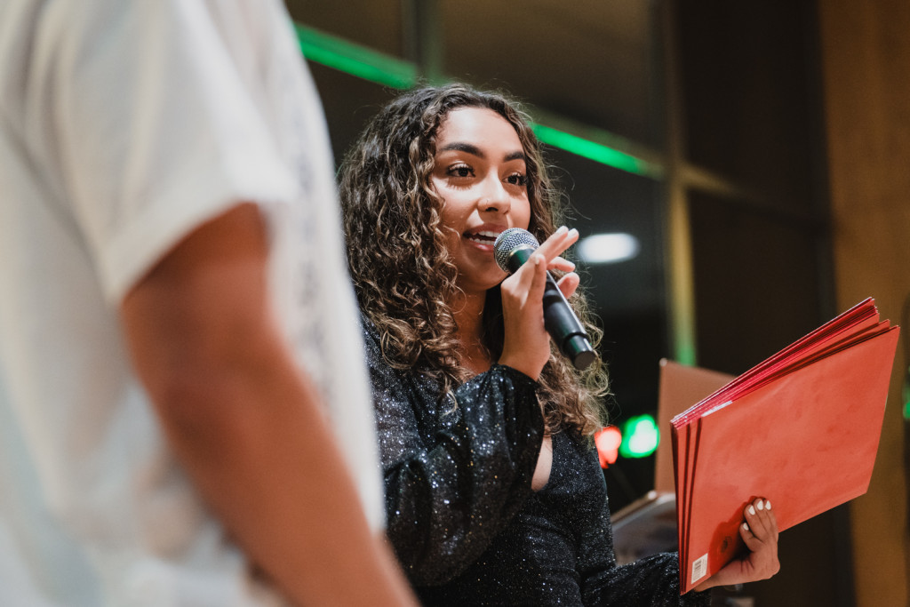 A woman holding red folders talks into a microphone.