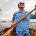 A man kneels in a birchbark canoe and paddles along a lake's shoreline.