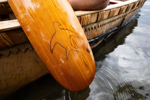 A close-up photo of detailing on a wooden paddle's blade shows a carving of a thunderbird icon. The paddle is held up from the water alongside the hull of the canoe.