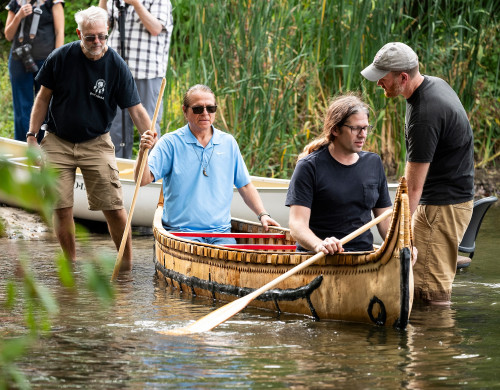 On a warmer day than the one of the canoe's first launch, Two men hold the ends of the canoe steady as two men sit in the canoe, ready to push off from shore.