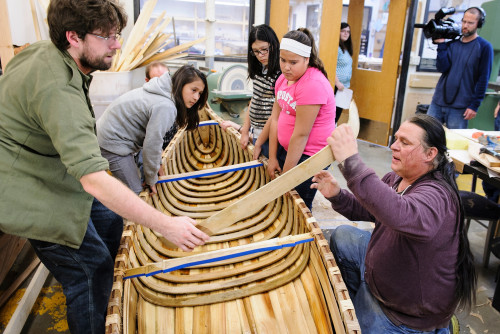 In a woodworking shop, wo men holding either end of a piece of birch lath over a half-finished birchbark canoe as they speak to a group of three sixth-grade students.