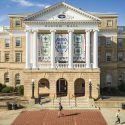 A view of the front facade of Bascom Hall taken from a drone. On a sunny day, a few people walk across the brick and concrete path in front of the building. The four panels of the Seed by Seed banner hangs between tall, white columns above the building's main entrance. The banner has been printed with a texture resembling beadwork and contains symbols and colors representing traditions of the Ho-Chunk Nation. Four green rings represent the four lakes of Teejop, the name the Ho-Chunk give the land now occupied by UW–Madison. Inside each ring, square patches in light blue, dark blue yellow and red represent the reflections of light on the water at different times of day. Two large pink triangles on either side of the banner represent flowers, with green stems and triangular leaves leading to the center panel. On the center panel, a large diamond made of small blue triangles frames a blue thunderbird, which is flanked by two red, abstract W's, representing UW–Madison. Below the thunderbird are two green water spirits, which resemble four-legged animals with very long tails. Below the water spirits are six light blue triangles representing water. Above and below the large diamond frame are bursts of yellow beading, representing the sun. Along the bottom border of the banners are stylized animal symbols of the twelve clans of the Ho-Chunk Nation, and beneath each animal is a traditional Ho-Chunk flower motif in blue and green.