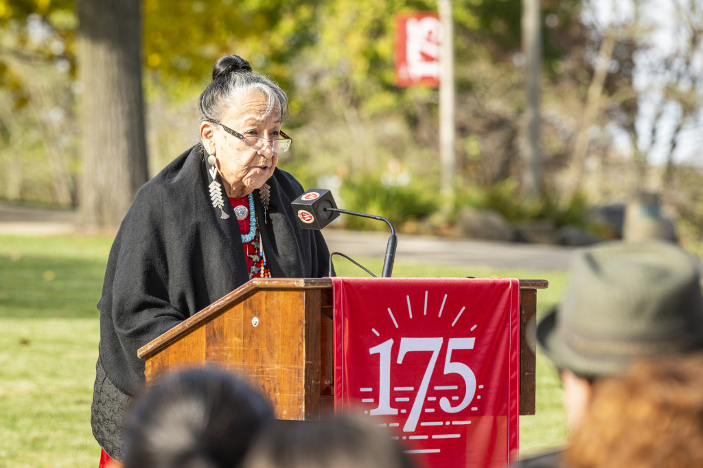 A woman wearing several beaded necklaces and pendants speaks from the podium.