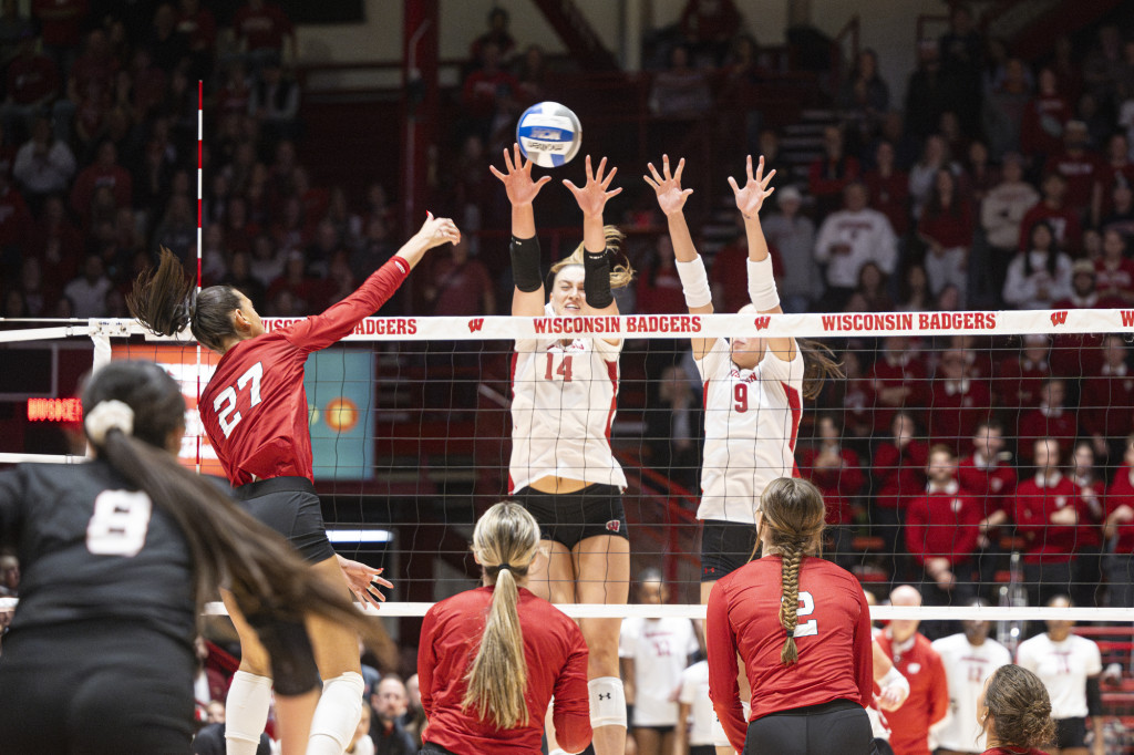 Two players reach high over the net, arms up, to block the ball.