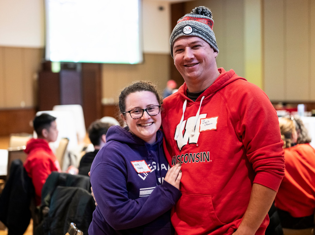 A woman wearing purple and a man wearing red stand with arms around each other.