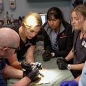 A group of veterinary medicine students and instructors wearing scrubs gather around an operating table.