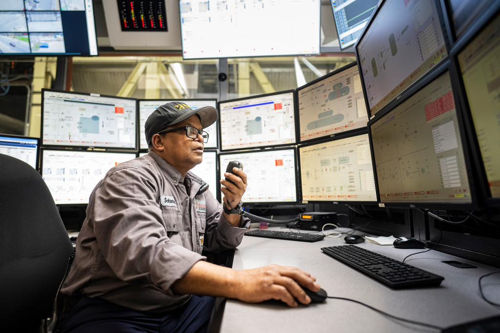 A man sits at a bank of monitors stacked two high and six wide. More monitors are mounted from the ceiling above him. He's speaking into a walkie talkie held in his left hand while he moves a computer mouse with his right.