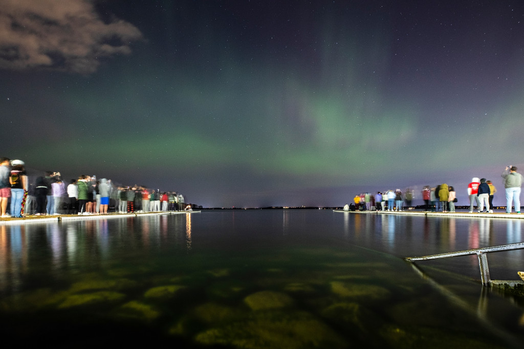 A starry night sky is lit by shades of green and purple as the aurora borealis shines down on Lake Mendota. Students taking in the view stand on piers extending out into the lake.