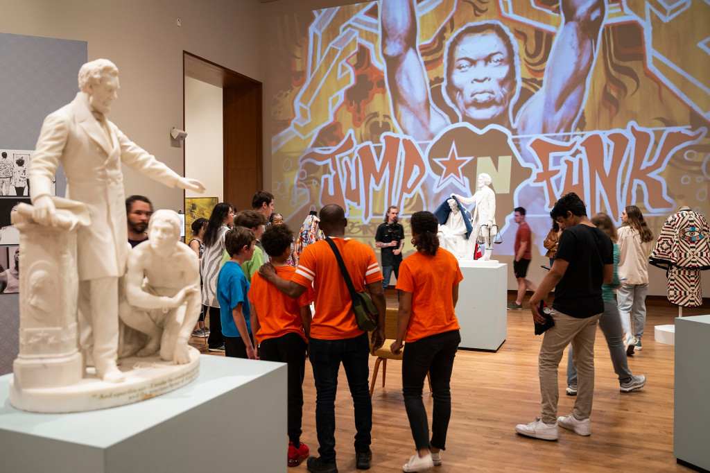 In an art gallery space, visitors to the Reemancipation exhibition take in two statues standing in juxtaposition across the floor from one another. In the first, Abraham Lincoln stands at a podium and reaches down to a crouched enslaved person whom he has just declared emancipated. The second, newer statue shows Fredrick Douglas standing, holding the scales of justice in his left hand as he pulls back a quilt from the head of a seated Abraham Lincoln. The wall of the gallery is covered floor-to-ceiling with a projection of a street-art-style mural showing a Black man with his arms overhead and the words Jump n Funk along the bottom.