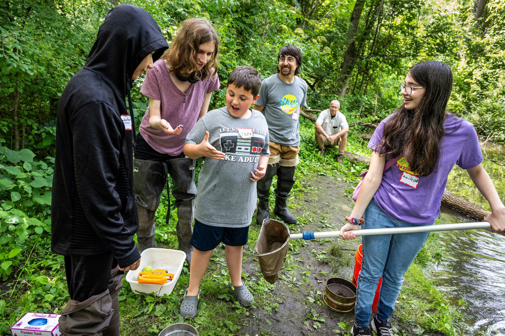 A group of four teenagers stand in the woods along the bank of a creek while their two instructors look on behind them. One of the teenagers is holding a long pole with a net on the end. She and two of her peers look on as a third student looks in wonder at what's in his hand.