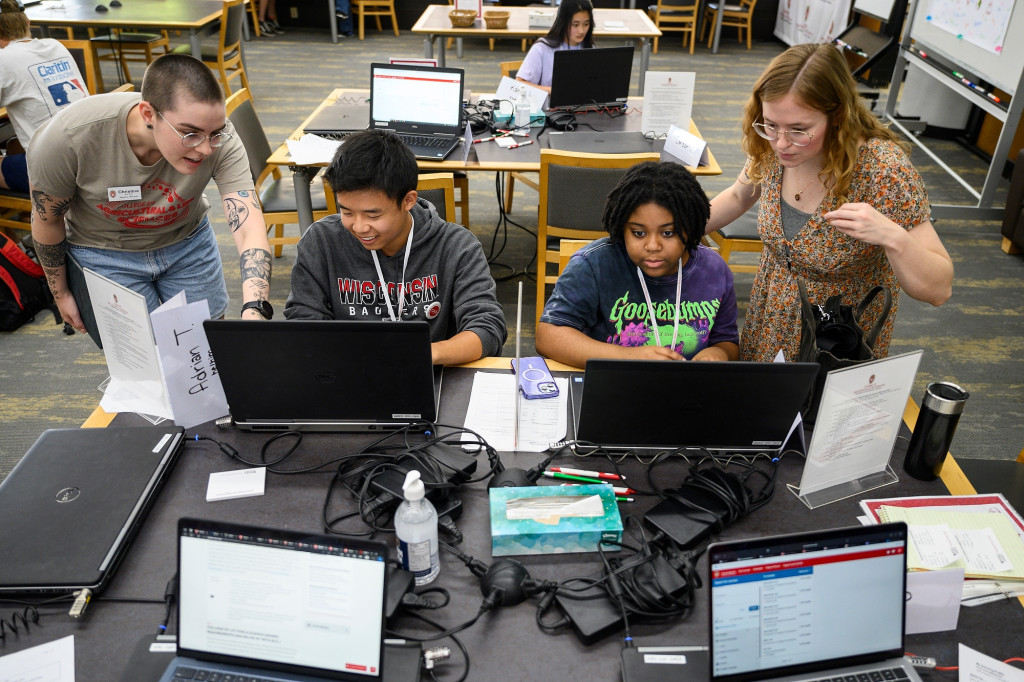 At a library table, two incoming first-year students sit at laptop computers while older students look over their shoulders to help them register for classes.