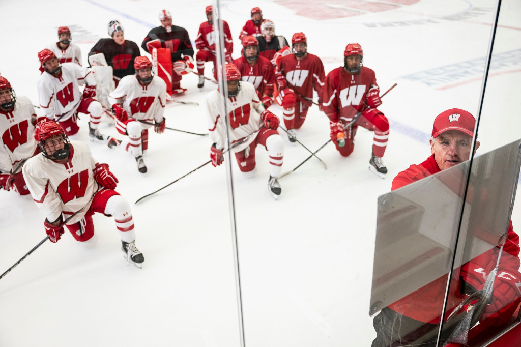 In a photo taken from above, the UW women's hockey team takes a knee on the ice as they listen to head coach Mark Johnson while he draws on a whiteboard.