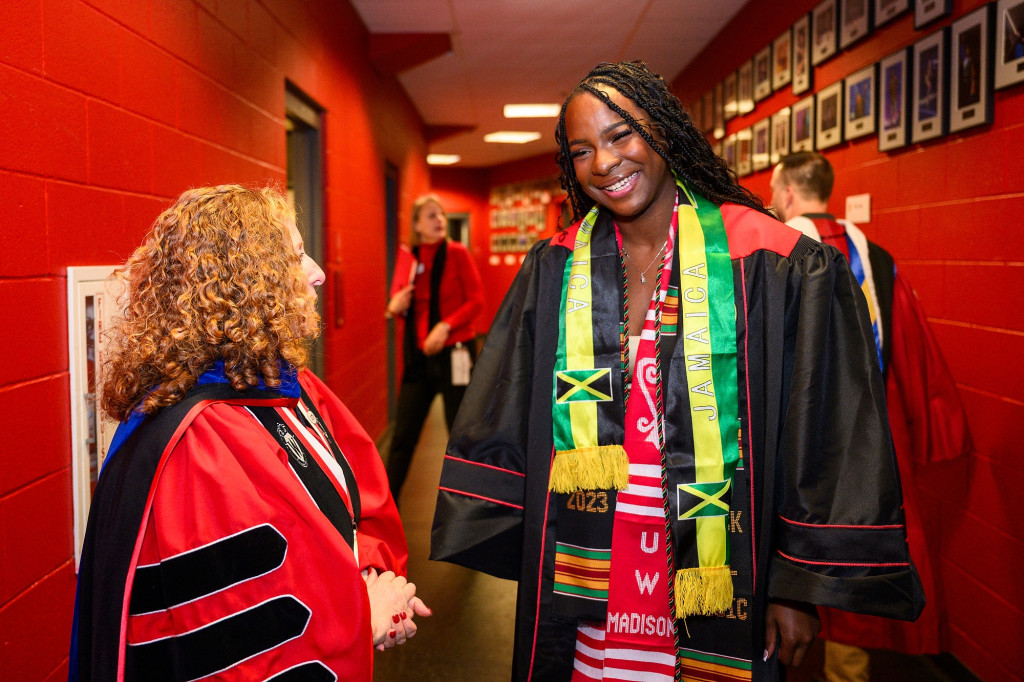 Two women chat in a hallway; both wear academic regalia.