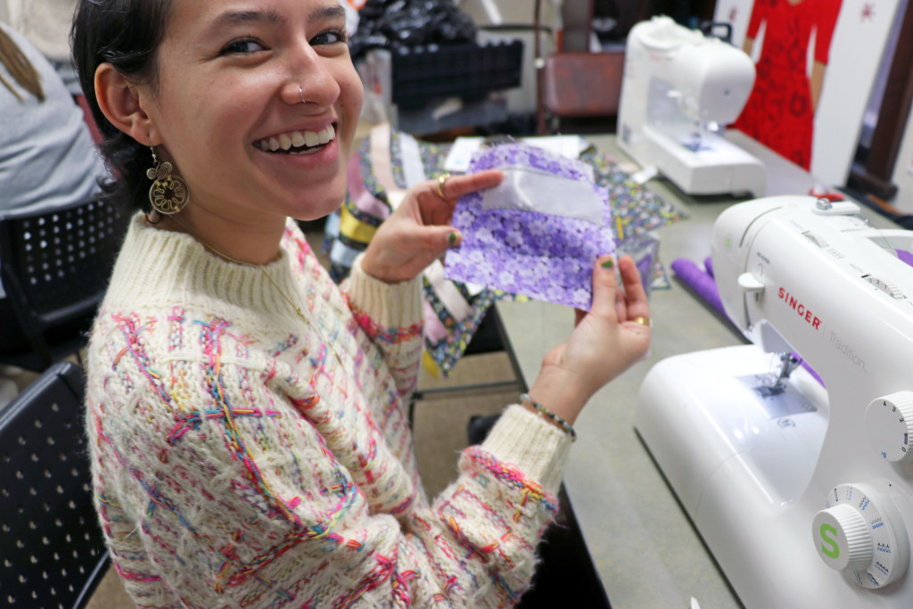 A student sits at a sewing machine and holds up a piece of fabric and matching ribbon.