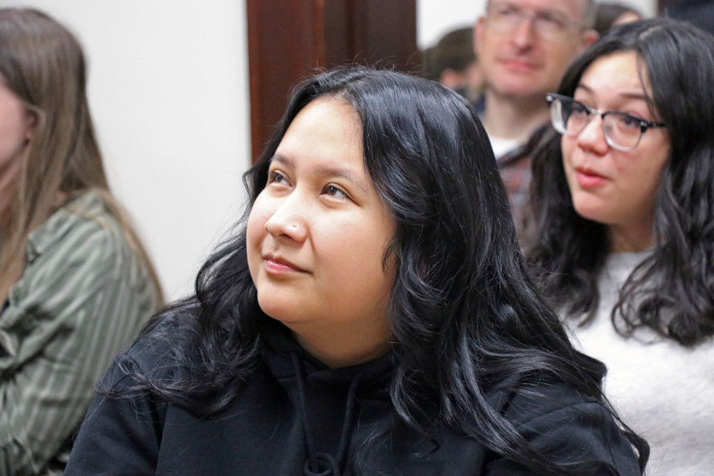 A close up of a young woman listening to someone speak at the head of a room.