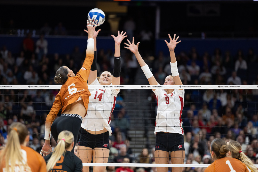 Two players jump in the air, arms raised, and block the ball.