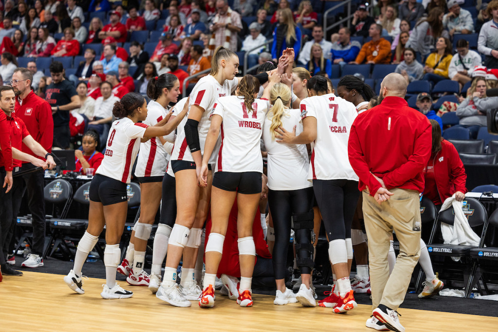 Players on the UW women's volleyball team huddle on the sidelines during the game, with the crowd-filled stadium in the background.