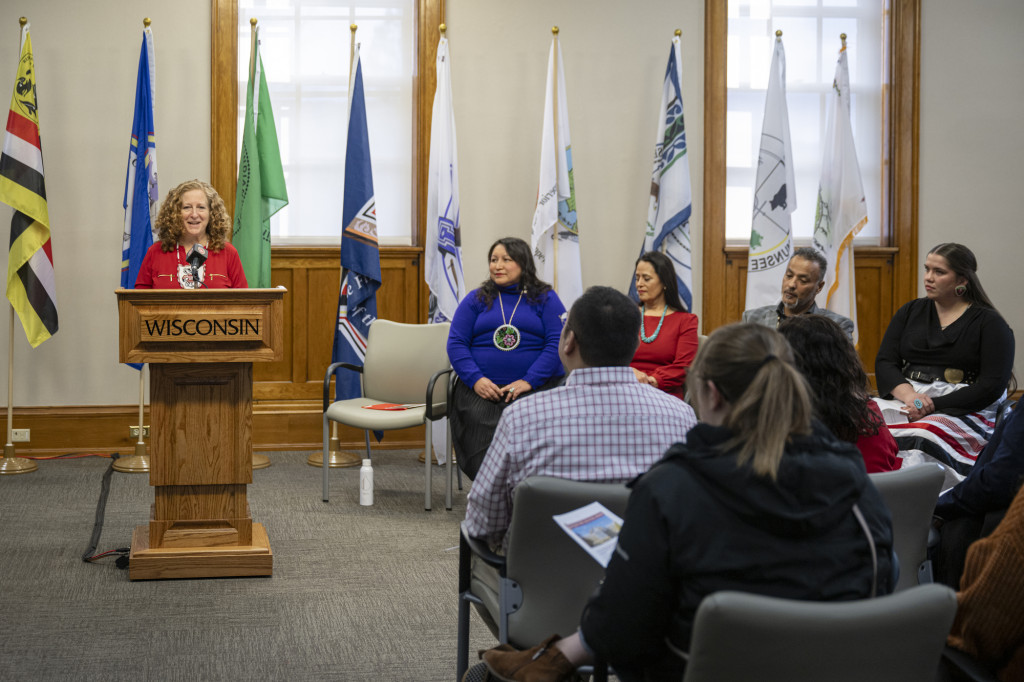 A woman talks at a podium while several others sit in chairs near her listening.