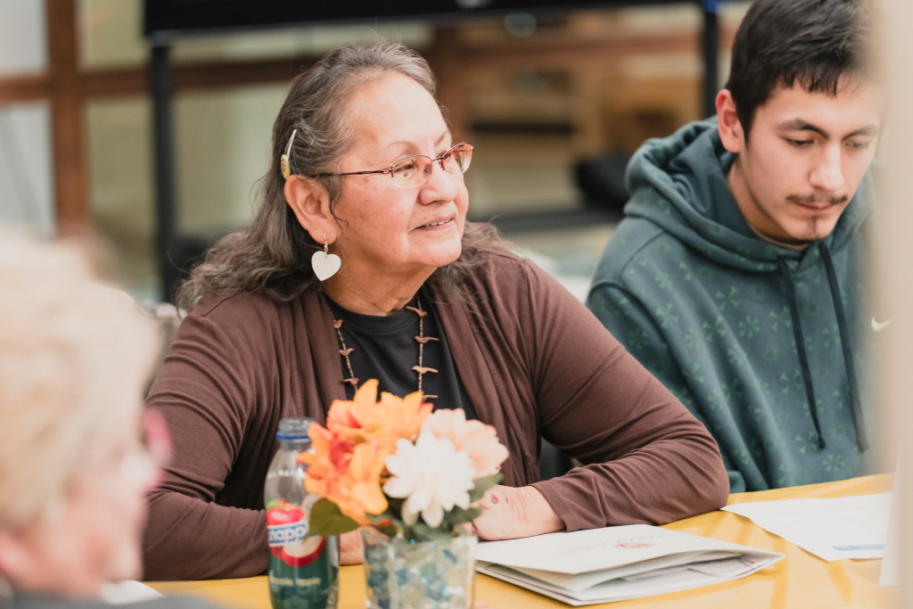 An older woman sits quiet at a table with arms folded while listening to a speaker.