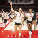 Volleyball players run off the court while smiling and yelling and raising their hands.