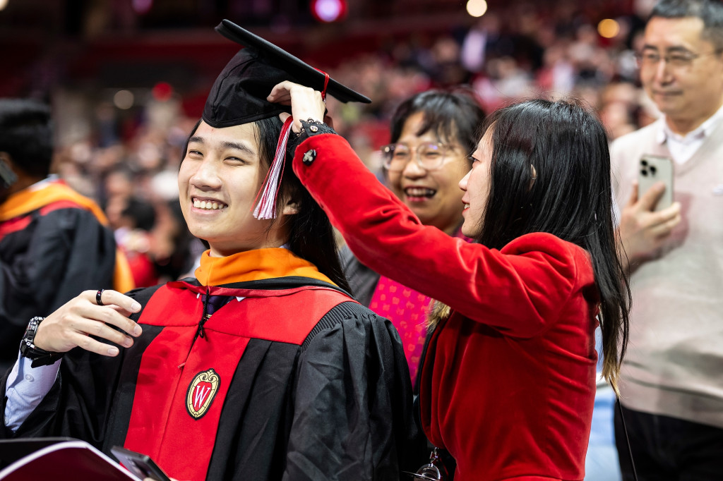 A man wearing a commencement robe and a mortarboard gets help adjusting his clothes.