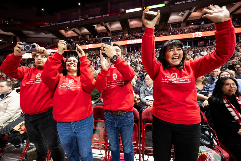 A group of people wearing red sweaters and standing in the Kohl Center stand and cheer.