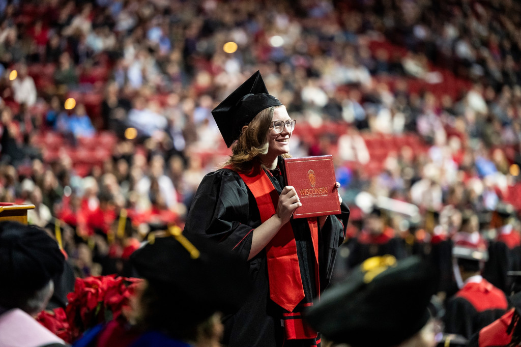 A woman holds up a diploma as she walks across a stage; other graduates are in the background.