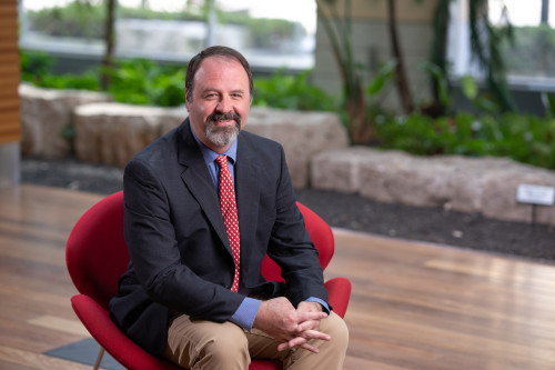 A photo of Jason Fletcher wearing a sports coat and tie, sitting in a red chair in the atrium of the Discovery Building on the UW–Madison campus.