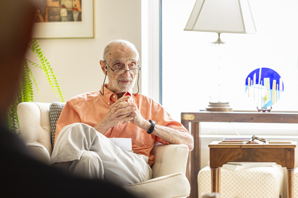 A photo of Bernard Cohen as an older man seated in an armchair in his brightly lit living room. He has his hands clasped together as he speaks to other people sitting off camera.