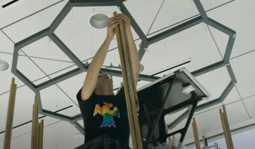 A photo of artist Chloris Lowe standing in the bucket of a crane hanging wooden rods from an installation in the shape of a porphyrin ring.