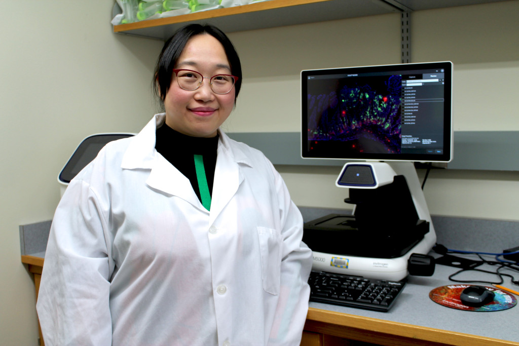 A woman in a lab coat stands in front of a computer screen.