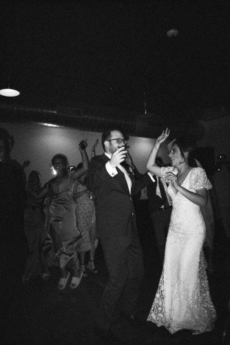 A black and white photo of Doug Olk in suit and tie and Rachel Olk-Lupini in her wedding dress with arms raised on the dance floor.