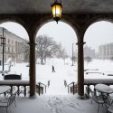 A person walks along snowy campus sidewalks; in the foreground are building arches.