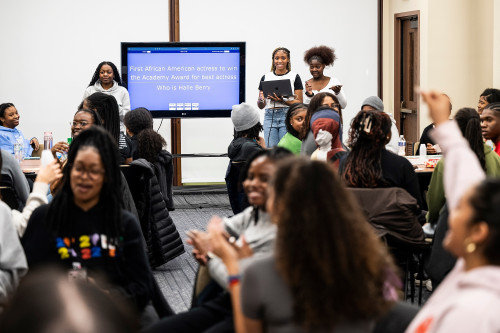 Three students stand at the front of a room quizzing participants in the style of Jeopardy.