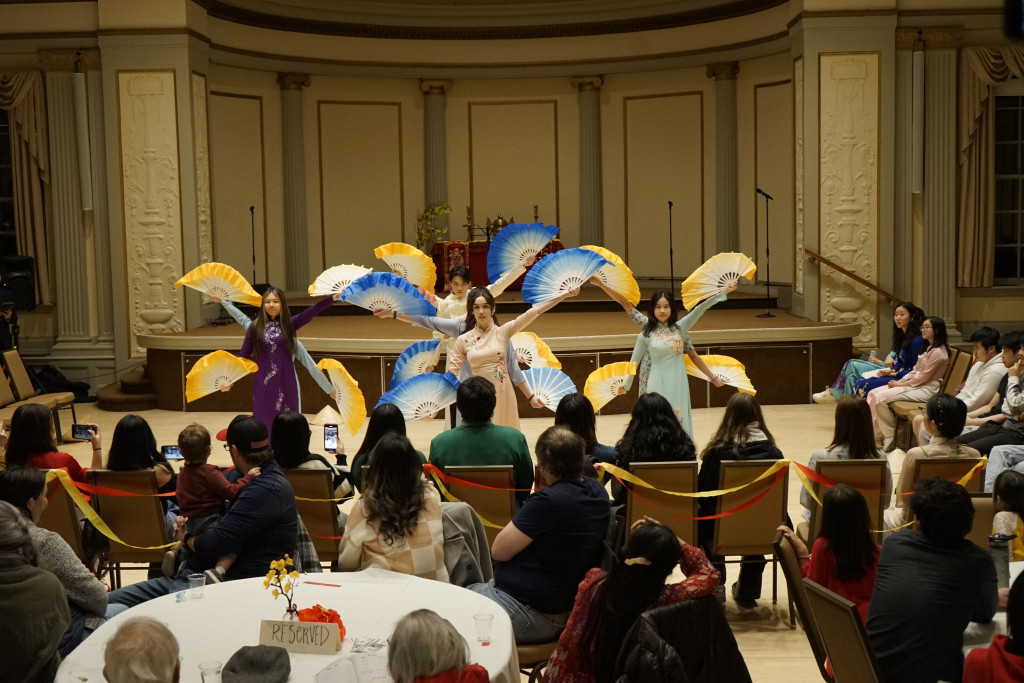 A group of performers move across a stage waving colorful blue and yellow fans in a choreographed dance.