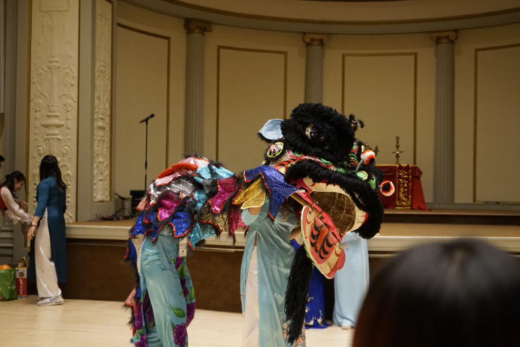 Two people wear a colorful Vietnamese lion costume over their heads and dance across a stage.