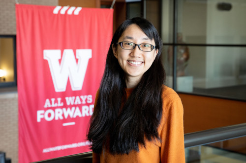 Photo of Ran Liu smiling and standing in front of a red and white banner that reads W All Ways Forward.