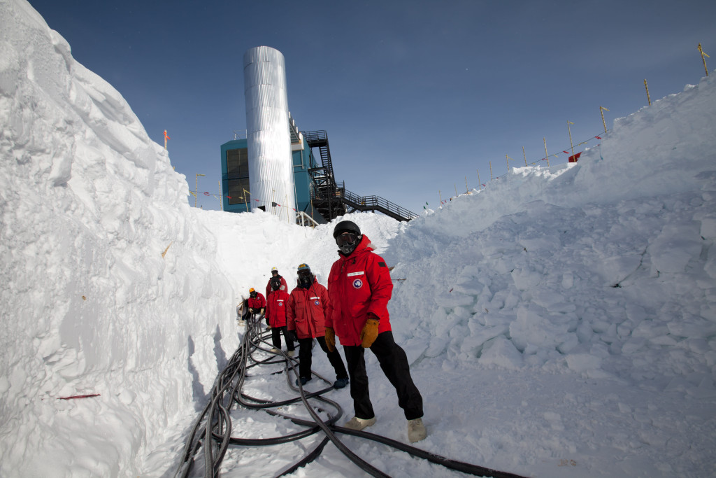 People wearing heavy clothes and red coats stand in a snow trench, with a network off thick wires at their feet.