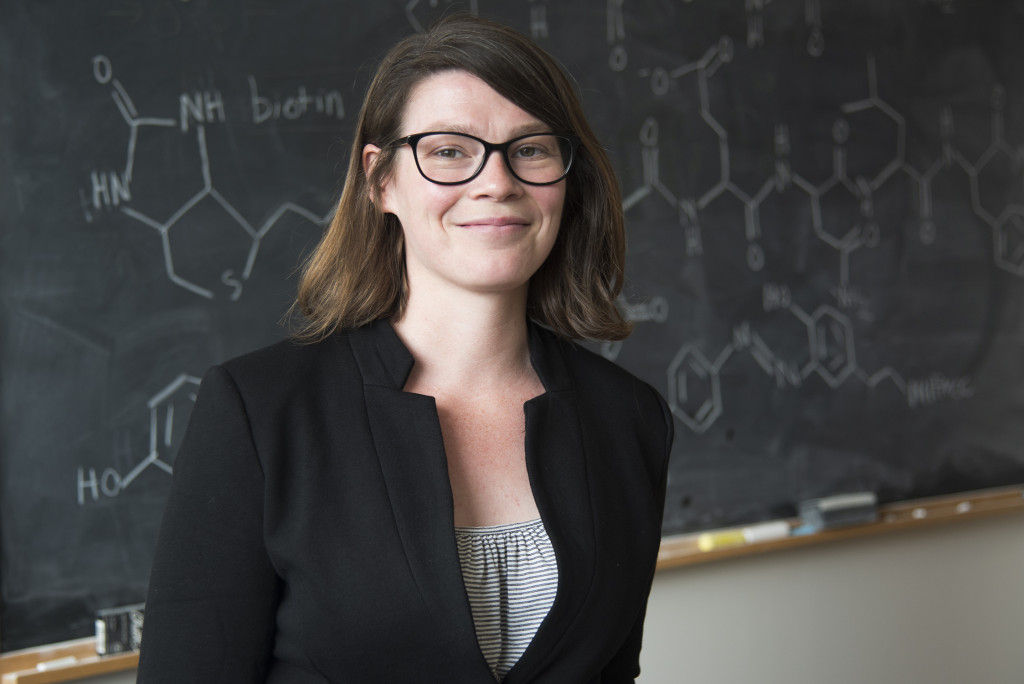 A photo portrait of Amy Weeks smiling. Behind her is a black chalkboard with drawings of chemical bonds.