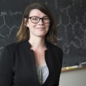 A photo portrait of Amy Weeks smiling. Behind her is a black chalkboard with drawings of chemical bonds.