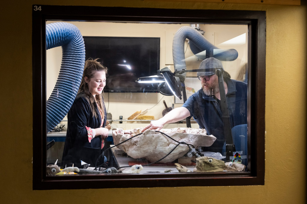 Two people, seen through an observation window, talk. A large stone is on the desk between them.