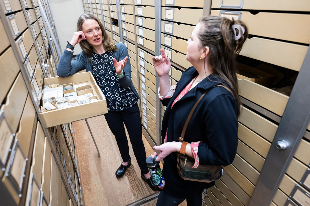 Two women stand between tall shelves, with one open drawer full of rocks between them, and talk.