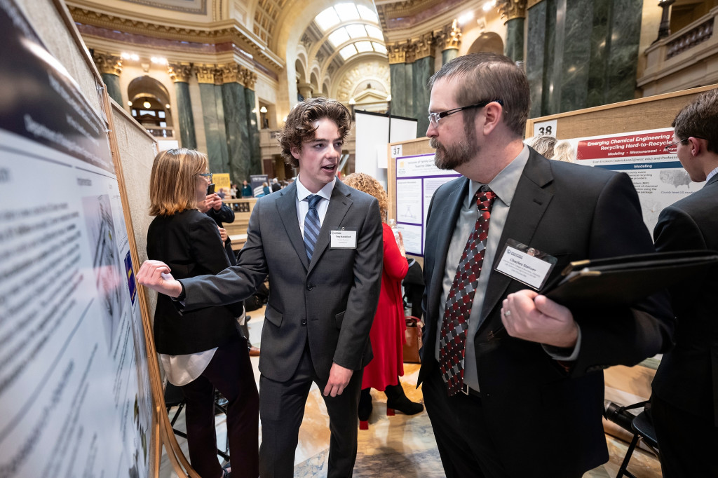 Two men wearing suits talk, one gestures at a poster board.